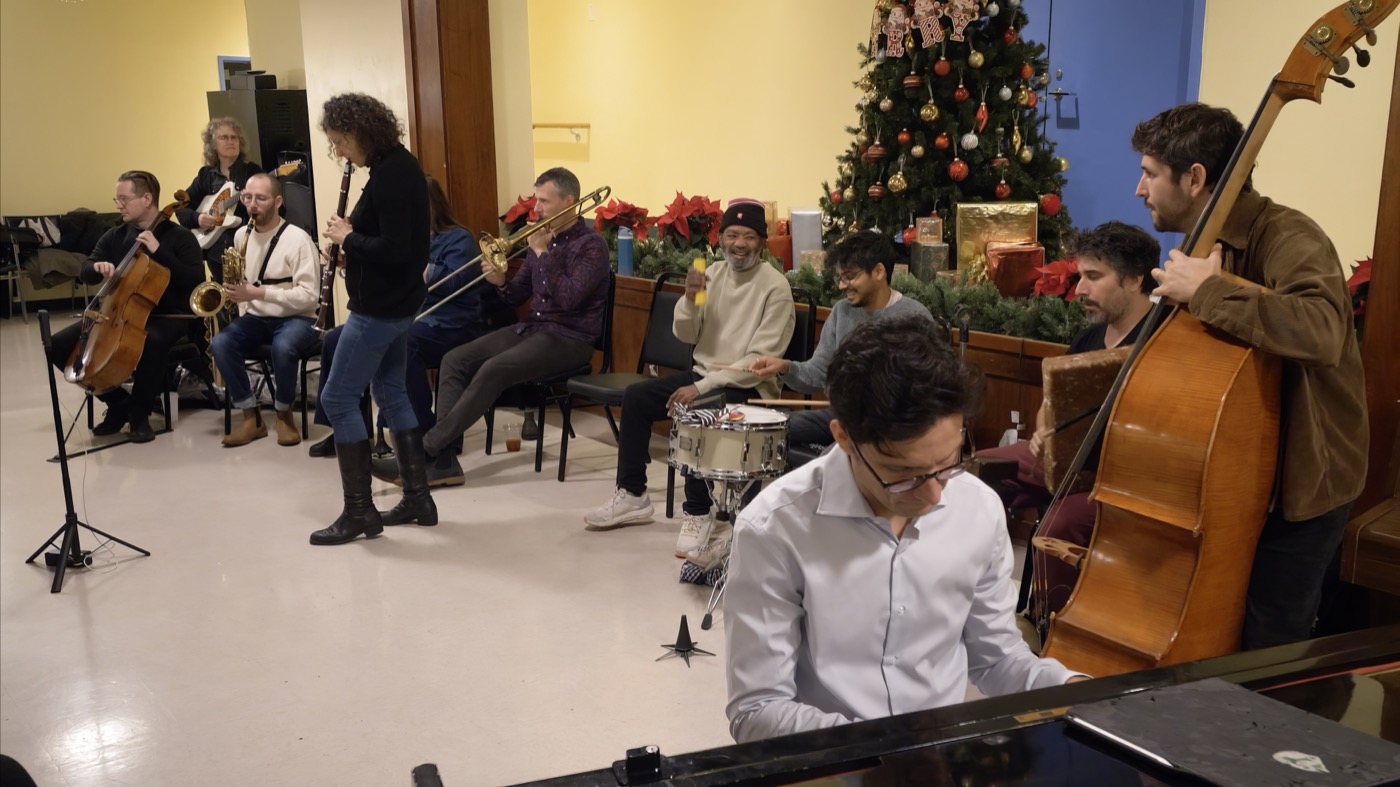 The ten-piece band jams in a circle in a holiday-decorated community room; a smiling community member plays a shaker beside the drummer.